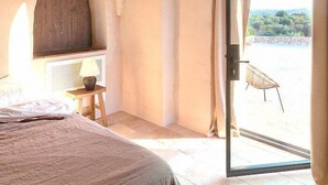 Un dormitorio con una cama, un banco de madera, una ventana con cortinas y una puerta que da a un espacio al aire libre.