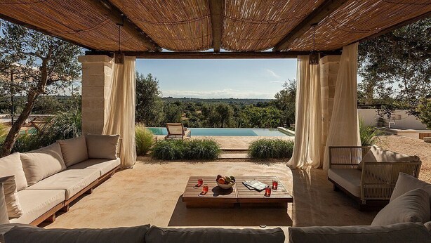 Un espacio al aire libre con vistas a la piscina, sofás acolchados, una mesa de madera con frutero y una pérgola.