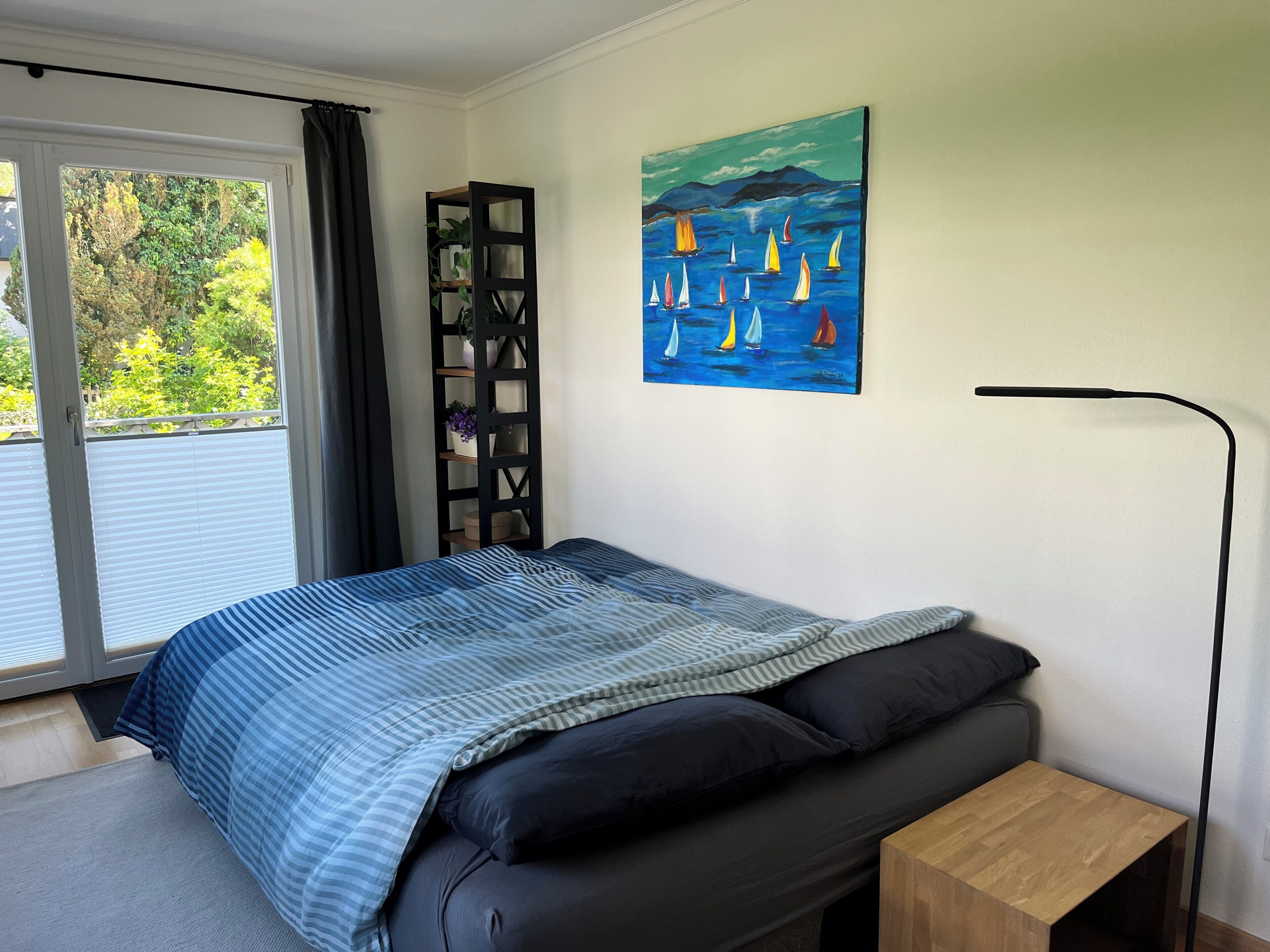A bedroom with a bed, a nightstand, a painting, a window with curtains, and a plant shelf.