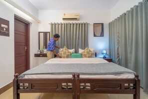 Habitación de hotel con cama de madera, mesita de noche con lámpara, aire acondicionado instalado en la pared y una puerta con interruptor de luz.