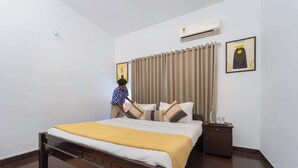 A neatly made bed with a yellow blanket, a wooden headboard, a nightstand with a bottle, and a wall-mounted air conditioner. - Alaya Stays 3BHK Riviera Residence Villa (Ambalapuzha)