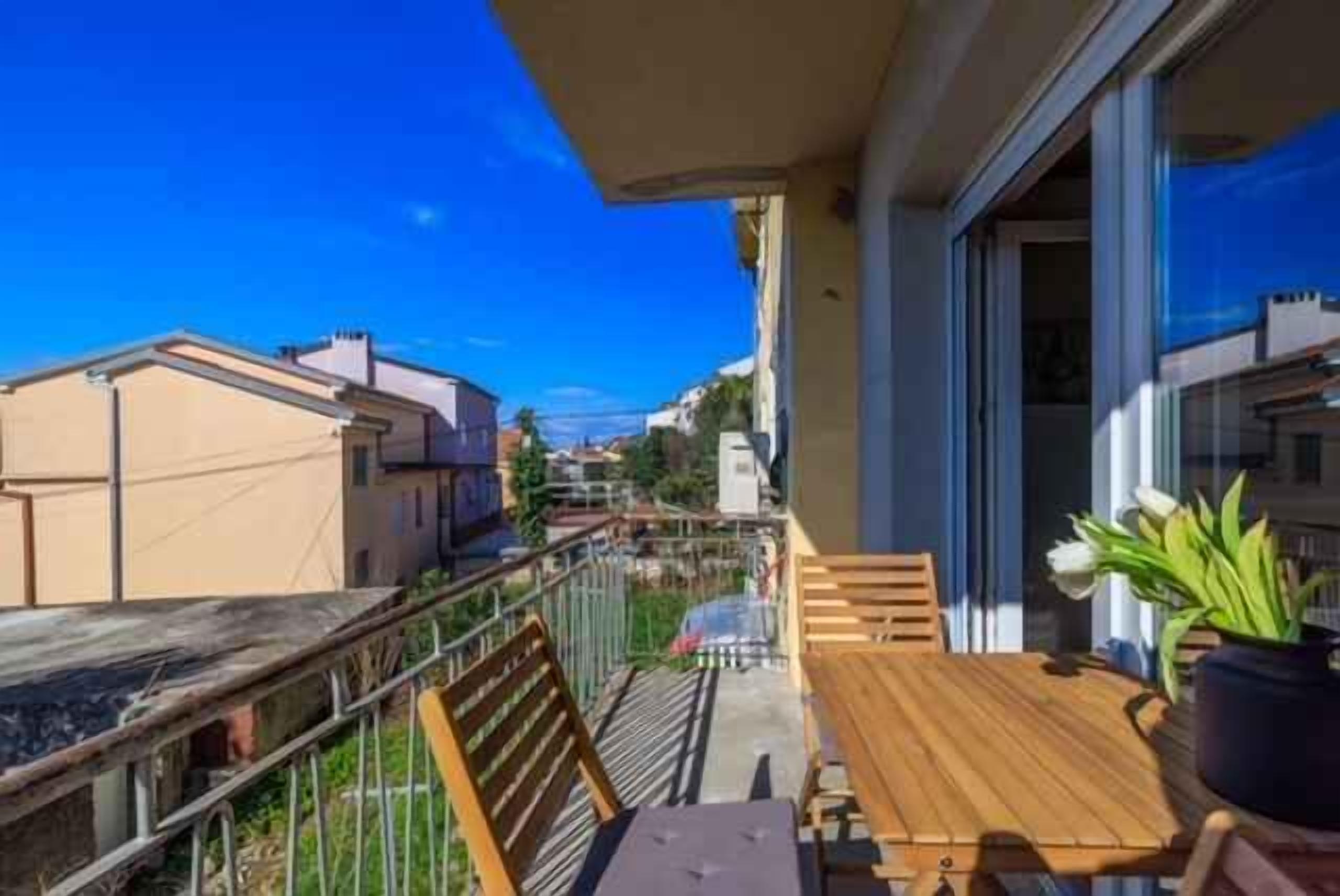 A balcony with a wooden table and chairs, a potted plant, and a view of neighboring buildings.