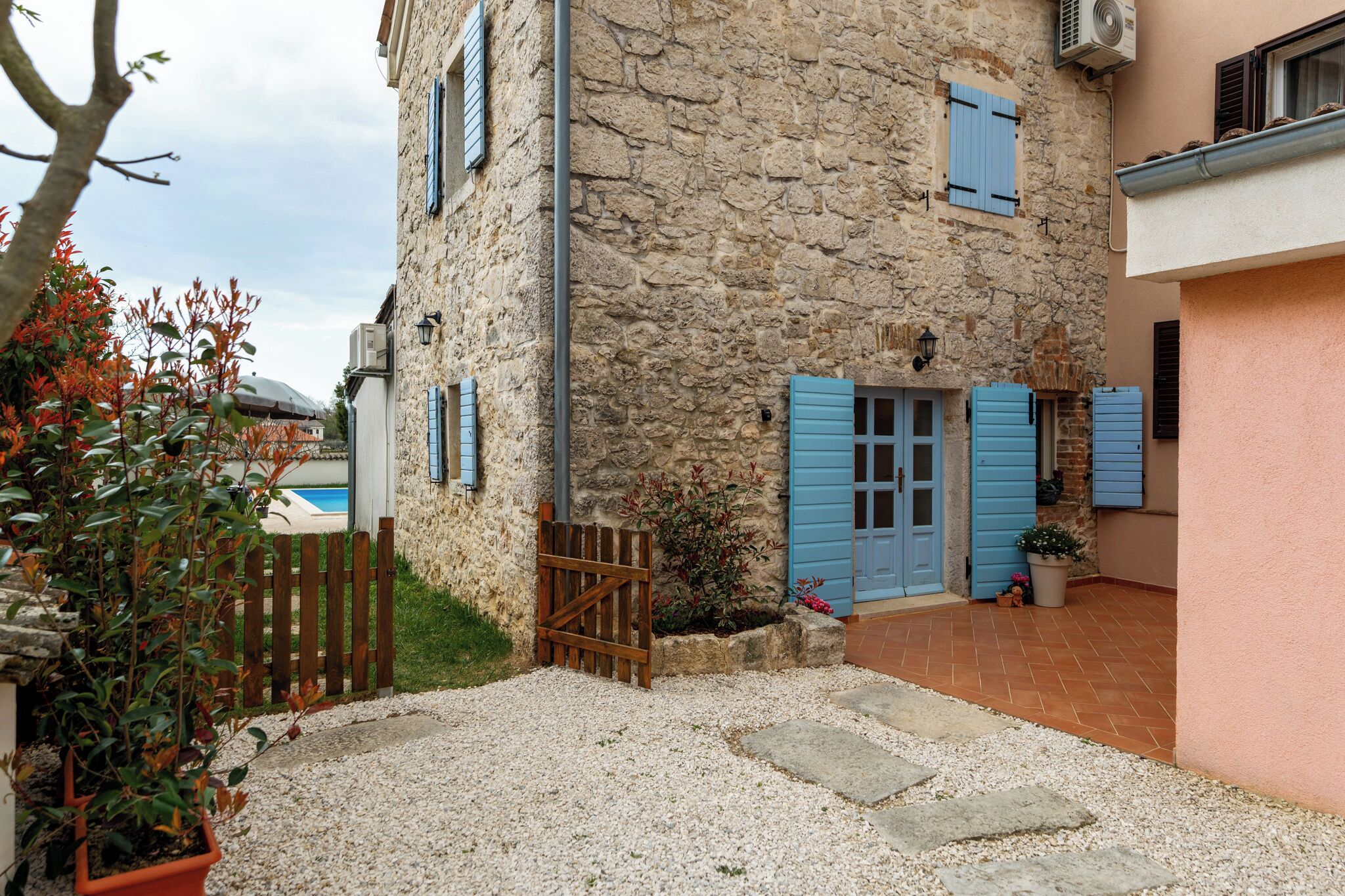 A stone building with blue shutters, a gravel driveway, and a pool in the background.