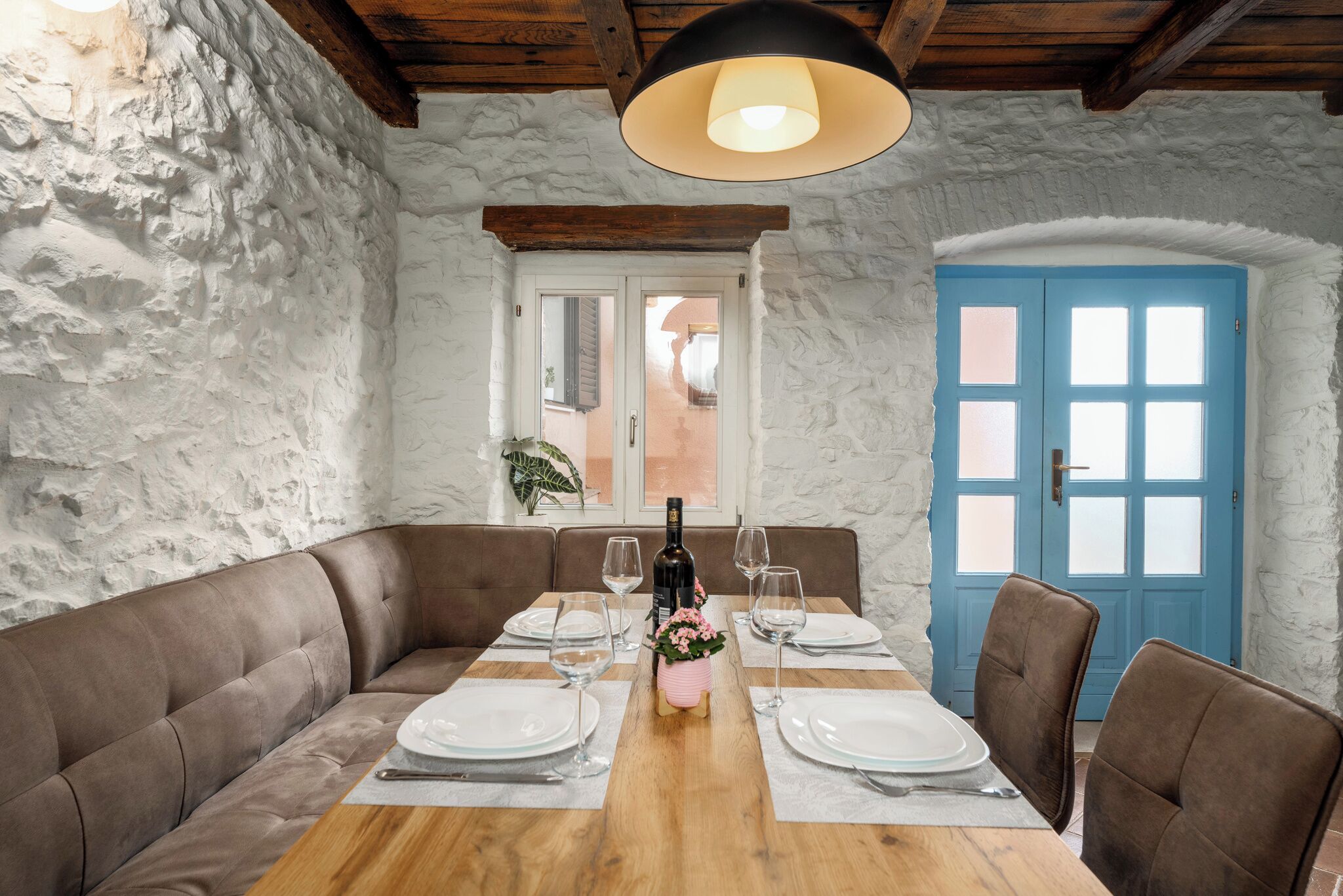 Un comedor con una mesa de madera, platos blancos y una botella de vino. Hay un banco acolchado marrón a lo largo de una pared y una puerta azul al fondo.