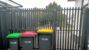 Tre svarta soptunnor med etiketter för återvinning, en svart metallgrind och ett asfalterat område.