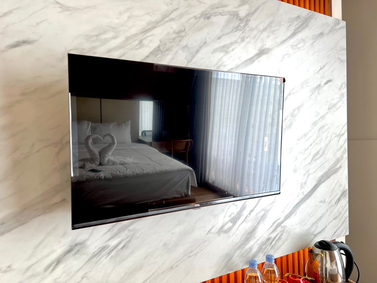 A flat-screen TV mounted on a marble wall, reflecting a room with a bed and curtains.