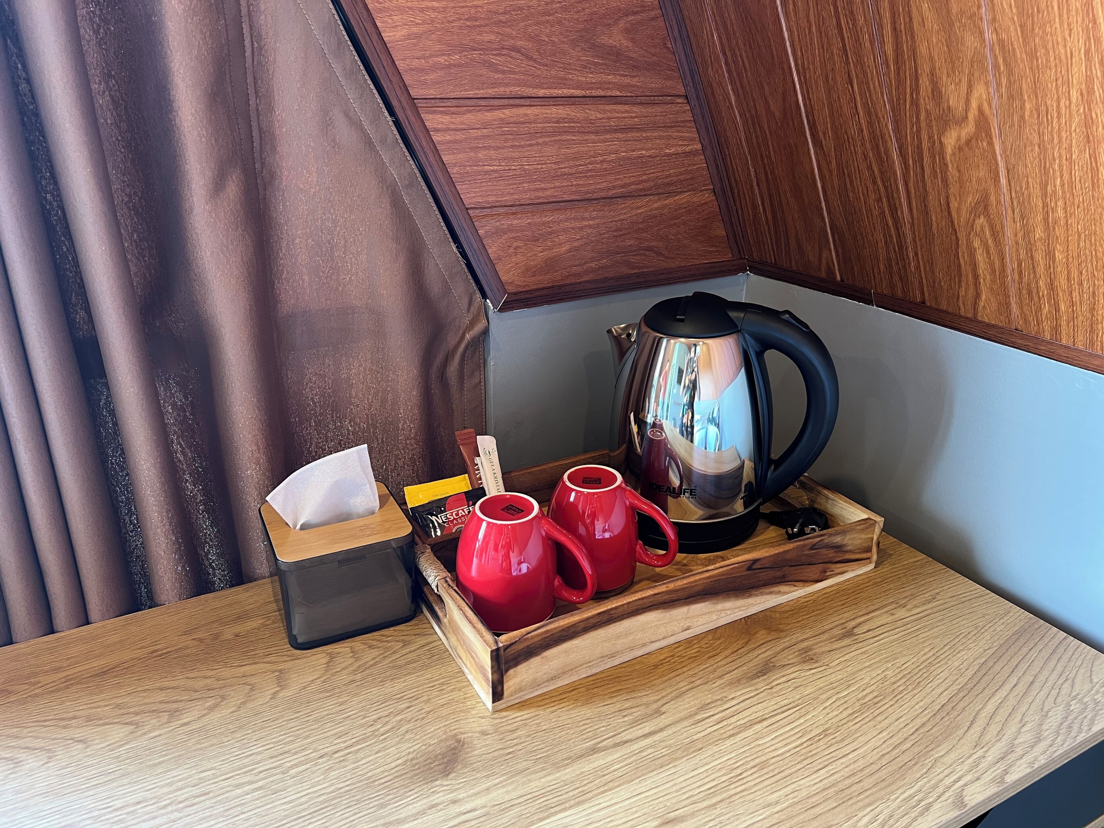 A wooden tray with a tissue box, a red mug, and a stainless steel kettle on a wooden table.