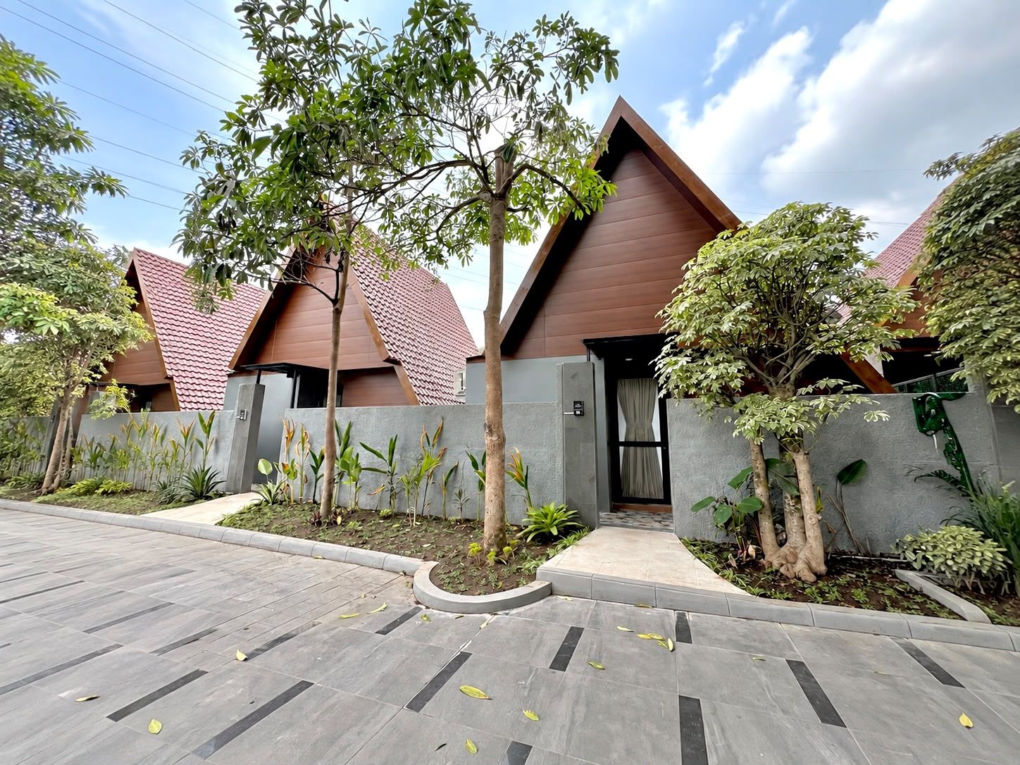 Um edifício moderno com um design triangular único no telhado, cercado por um jardim arborizado com diversas plantas e árvores.