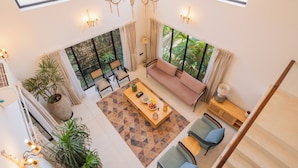 A spacious living area with a sofa, coffee table, and chairs, large windows with curtains, and a chandelier. - ELIVAAS Bogenvilla Luxe 3 BHK Villa (Anjuna)