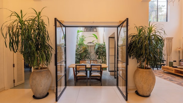 A dining area with a table and chairs, surrounded by large potted plants, and a view of the outdoor garden through a glass partition. - ELIVAAS Bogenvilla Luxe 3 BHK Villa (Anjuna)