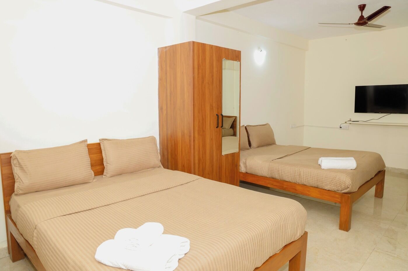 A room with two beds, a wooden wardrobe, a mirror, a ceiling fan, and a TV mounted on the wall.