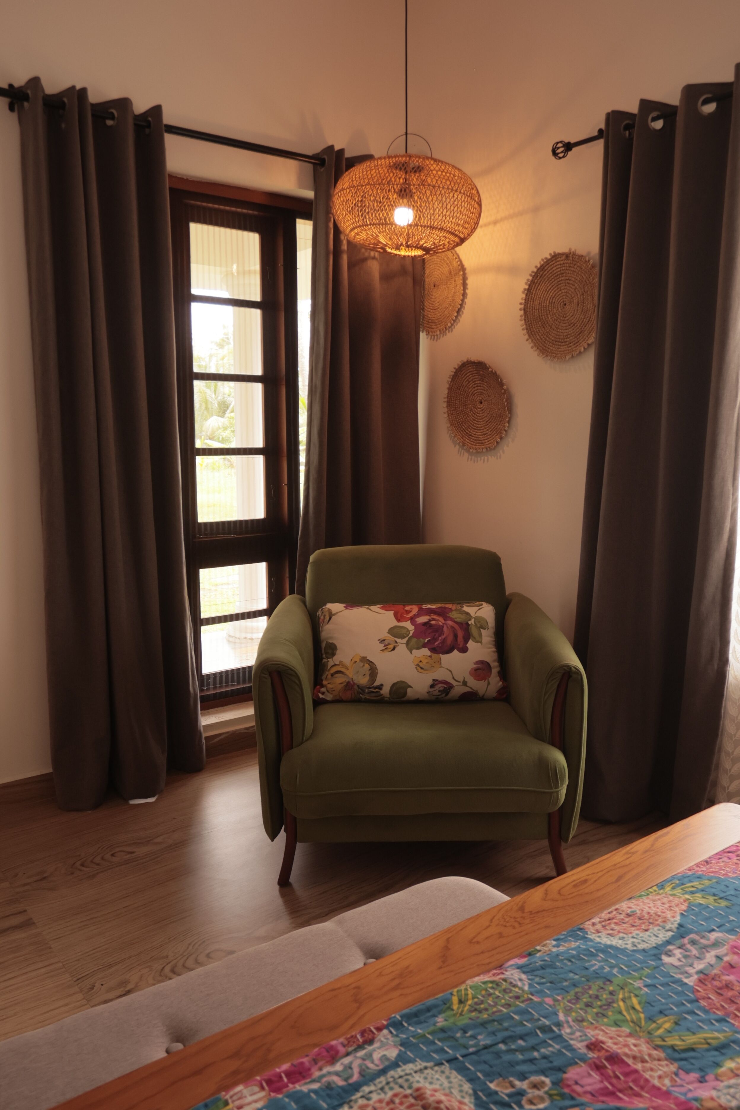 Ein Zimmer mit einem grünen Sessel, einer gewobenen Leuchte, einem gemusterten Teppich und Blick durch ein Fenster mit Vorhängen.