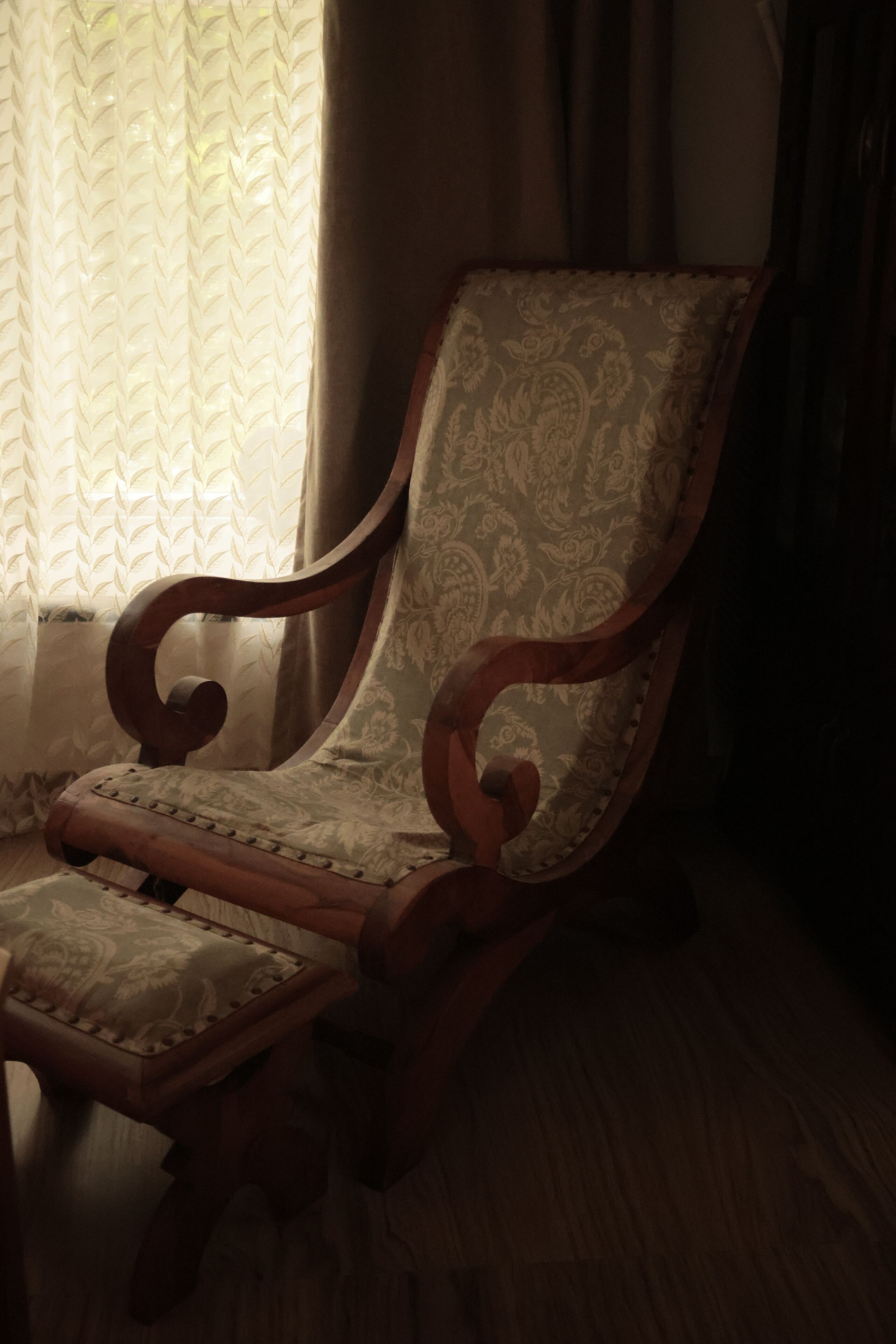 A wooden chair with a patterned cushion, positioned near a window with a patterned curtain.