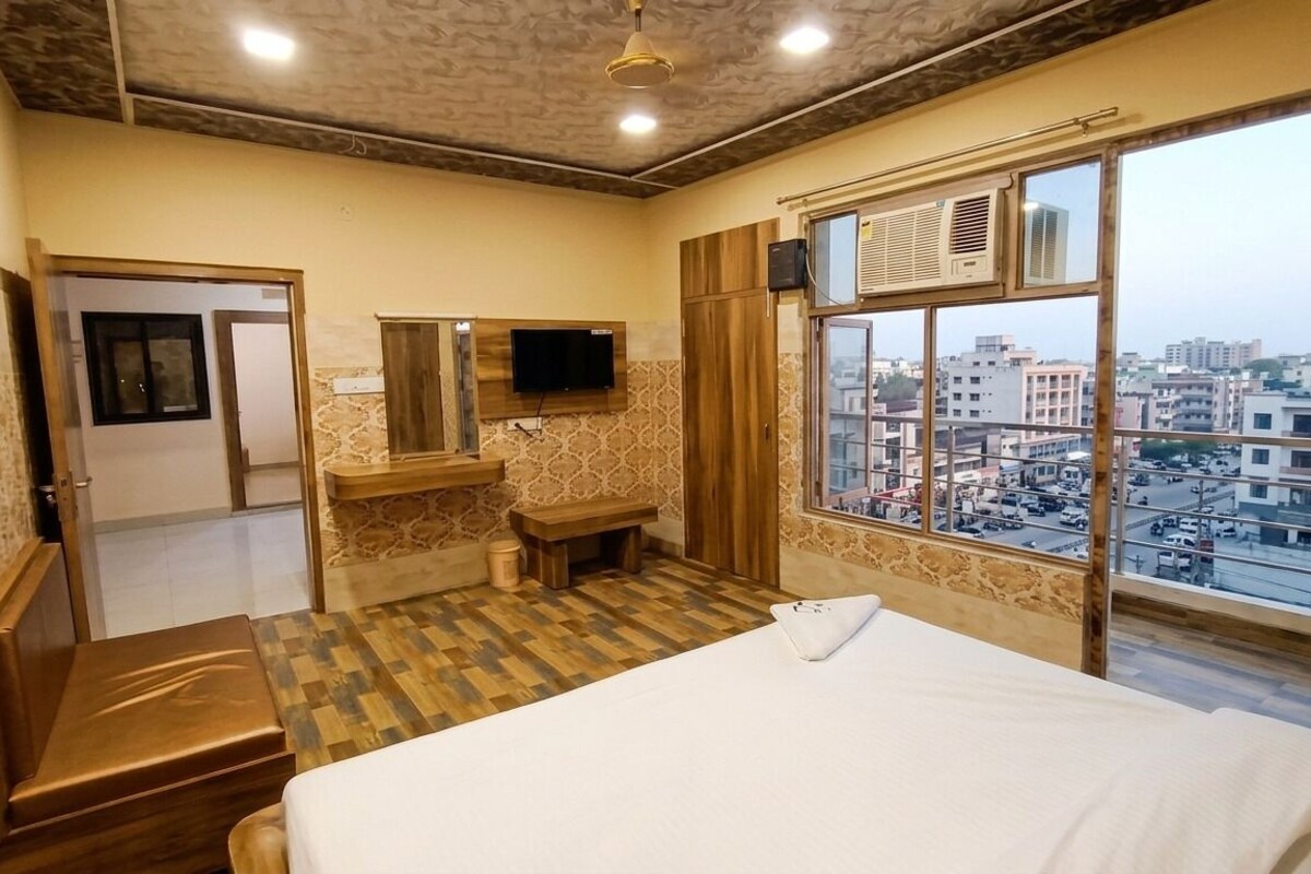 Een hotelkamer met een bed, een aan de muur bevestigde televisie, een raam met uitzicht over de stad, en een houten bankje.