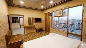 A hotel room with a bed, a TV mounted on the wall, a window with a city view, and a wooden bench. - Chhabra Hotel Kanpur (Kanpur)