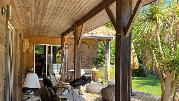 Terraza de madera con zona cubierta, mobiliario exterior y vista a un jardín con palmeras.