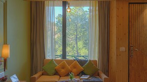 A cozy room with a wooden ceiling, a sofa with cushions, a small table with a lamp, and a window with curtains. - ELIVAAS 1BHK Pine View Resort  (Krishangarh)