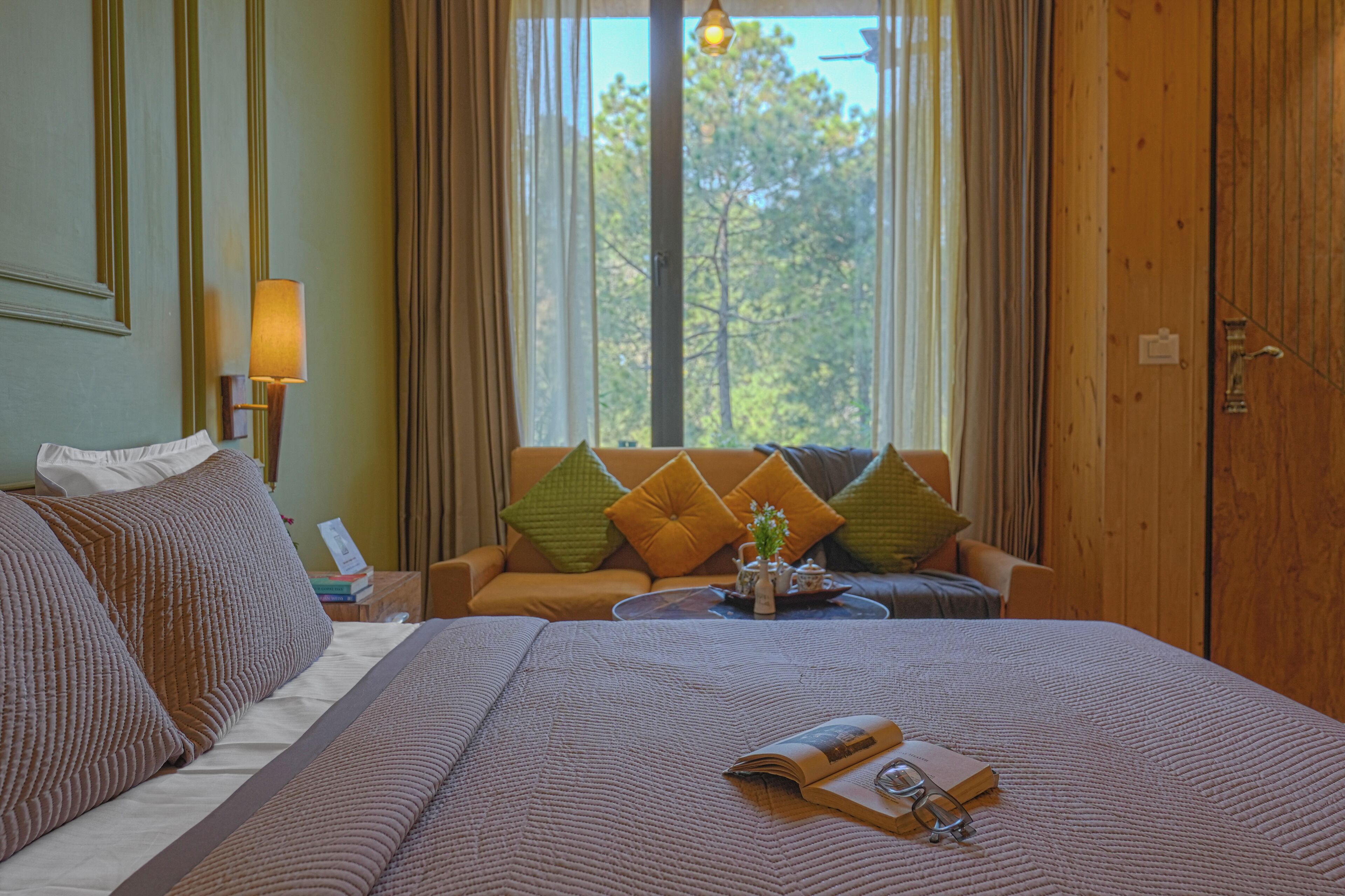 Habitación de hotel con una cama grande, un sofá, una mesa con lámpara y vistas a árboles por la ventana.