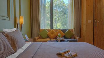 Habitación de hotel con una cama grande, un sofá, una mesa con lámpara y vistas a árboles por la ventana.