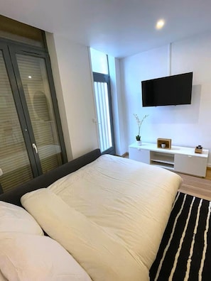 Un dormitorio moderno con una cama grande, un televisor montado en la pared, una mesita de noche con un jarrón y una puerta corredera de vidrio.