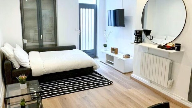A modern hotel room with a bed, a TV, a coffee machine, a mirror, and a seating area with a chair and a table.