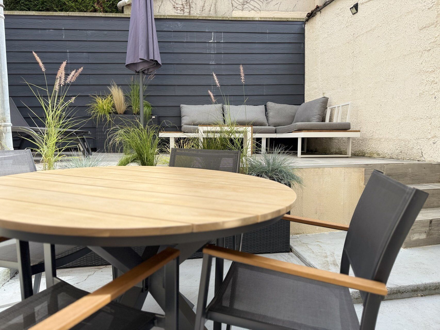 Una mesa redonda de madera con sillas, un sofá gris y plantas en macetas en un patio.
