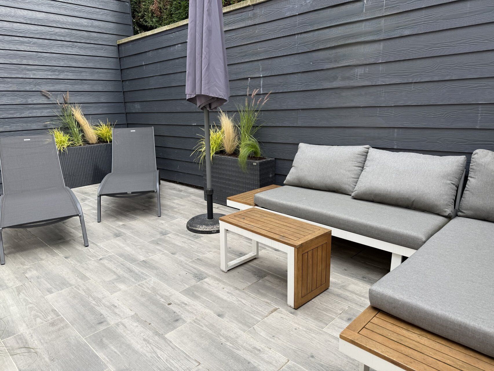 A modern outdoor seating area with a grey sofa, two chairs, a wooden table, and a potted plant.