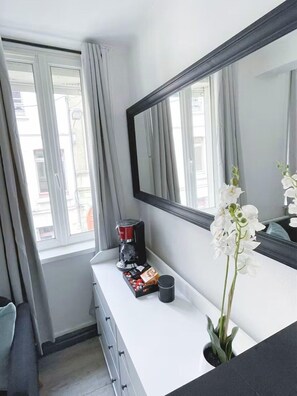 A hotel room with a large mirror, a coffee maker, a small plant, and a window with curtains. - The Refuge of the Squares (Arras)