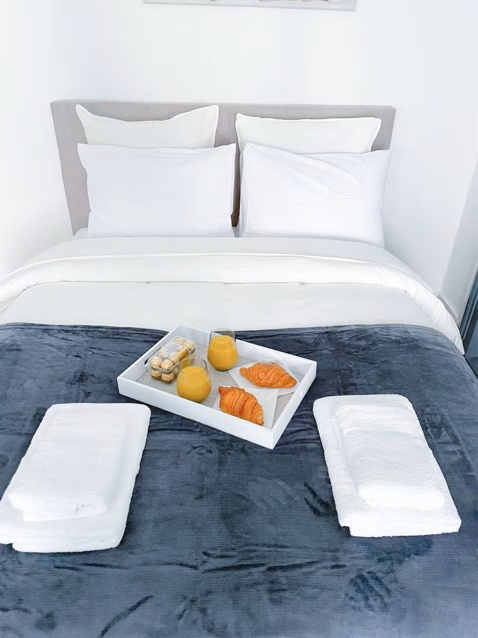 En snyggt bäddad säng med vita lakan, ett brickbord med croissanter, juice och en liten behållare, samt två hopvikta vita handdukar.
