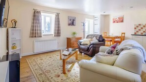 A living room with a sofa, a coffee table, a TV, a radiator, a window with curtains, a dining area with a table and chairs, and a kitchen area. - 1 bedroom accommodation in Llanfair Caereinion (Llanfair Caereinion)