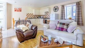 A living room with a brown leather chair, a white sofa, a glass coffee table, and a kitchen in the background. - 1 bedroom accommodation in Llanfair Caereinion (Llanfair Caereinion)