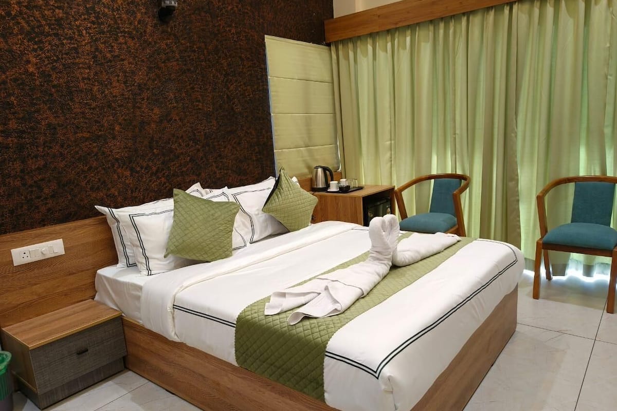 A neatly made bed with pillows and a blanket, a wooden bedside table with a kettle, a chair, and a green curtain.
