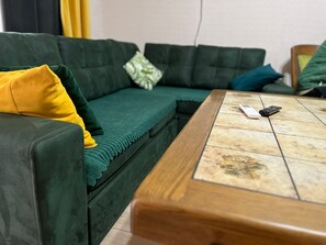 A green sofa with yellow and green cushions, a wooden coffee table with a remote control, and a patterned tile floor. -  Comfortable and Affordable apartment in Tbilisi (T'bilisi)