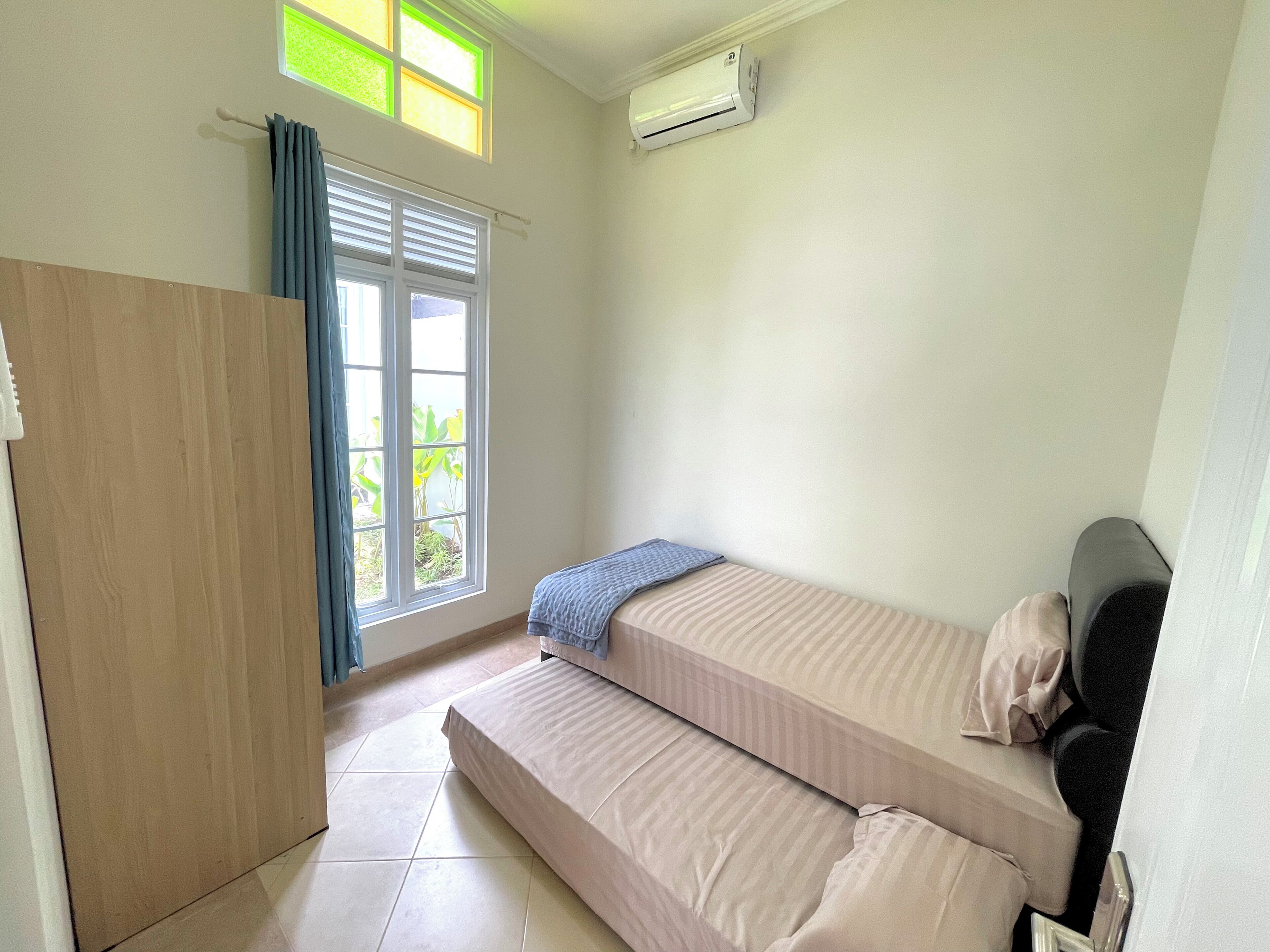 A small, clean room with a bed, a window with curtains, and an air conditioning unit.