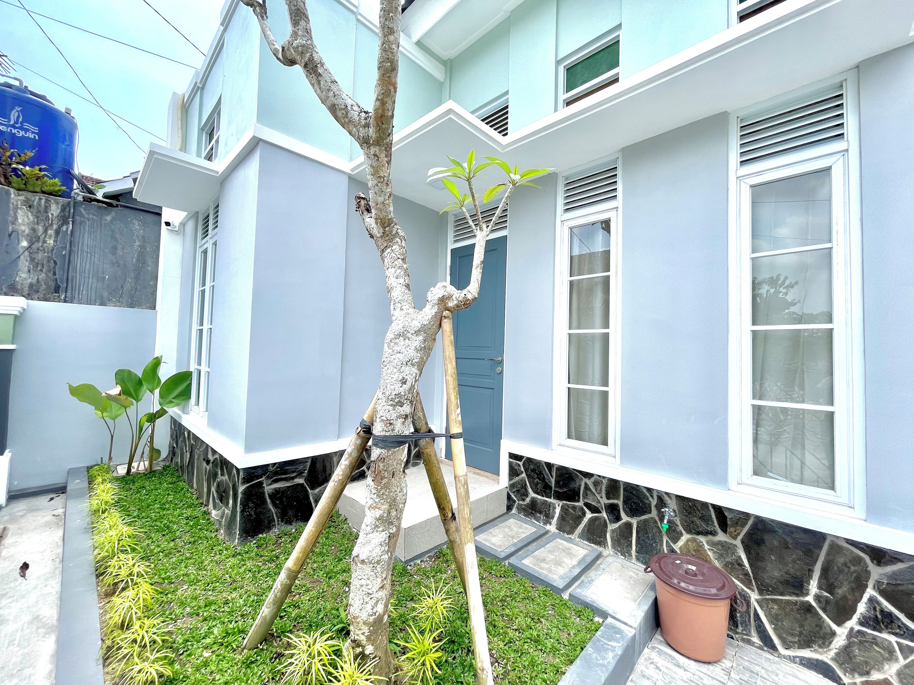 A residential building with a blue exterior, a tree in the front yard, and a potted plant on the porch.