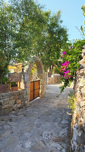 A stone pathway leading to a wooden gate with a stone archway, surrounded by greenery and a clear sky.