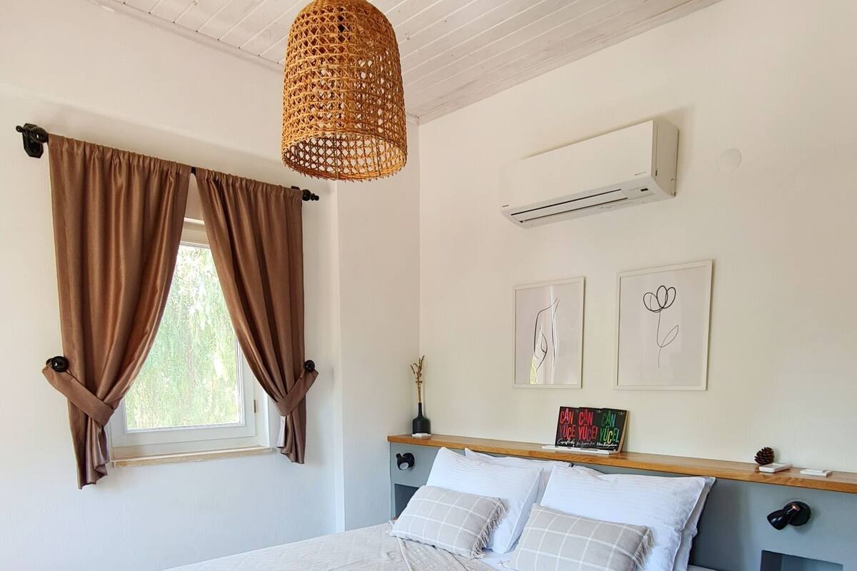 Una camera da letto con un letto, una lampada a sospensione, una finestra con tende, un condizionatore d'aria e un comodino con lampada.