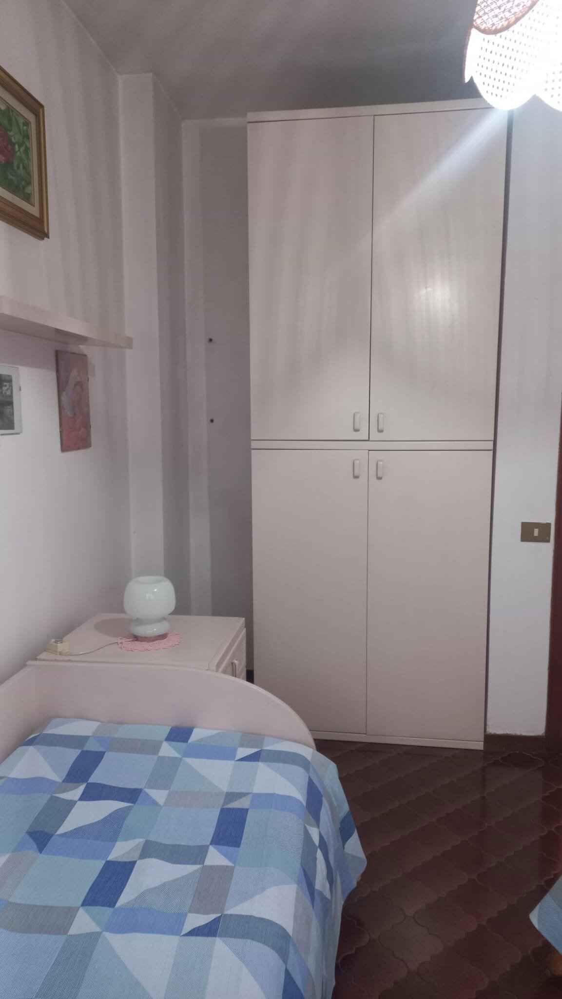 Ein Schlafzimmer mit einem Bett, einem Kleiderschrank, einem Nachttisch mit Lampe und einem an der Wand befestigten Regal.