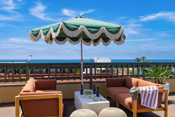 Ein Balkon mit einem grünen Sonnenschirm, zwei Sofas, einem kleinen Tisch mit Flaschen und Blick auf das Meer.