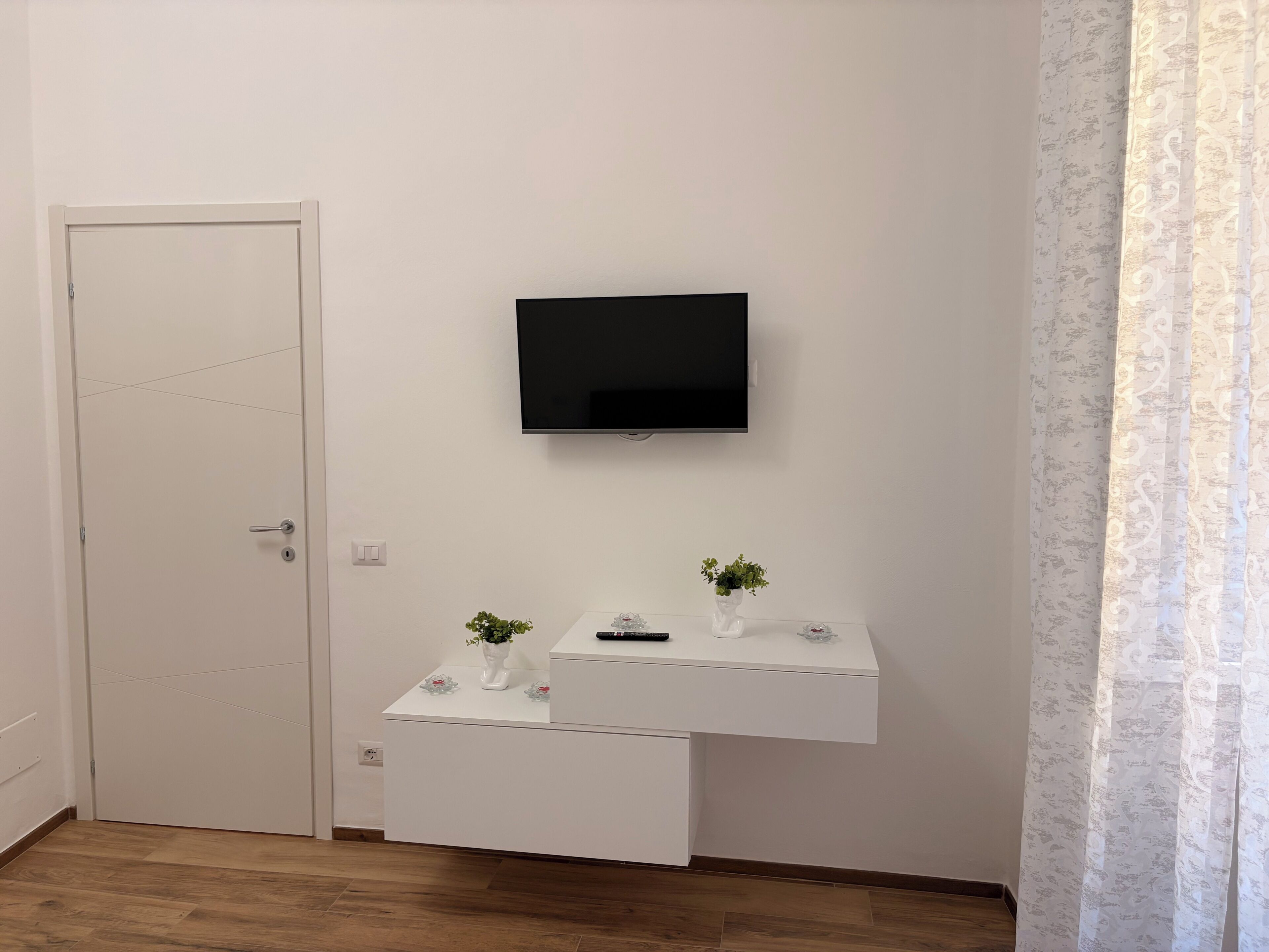 Huoneessa on valkoinen ovi, seinälle kiinnitetty televisio, pieni valkoinen työpöytä, jossa on kasvi, ja kuviollinen verho.