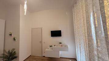 Una habitación con piso de madera, una puerta blanca, un televisor montado, una pequeña mesa blanca con una planta y una cortina estampada.