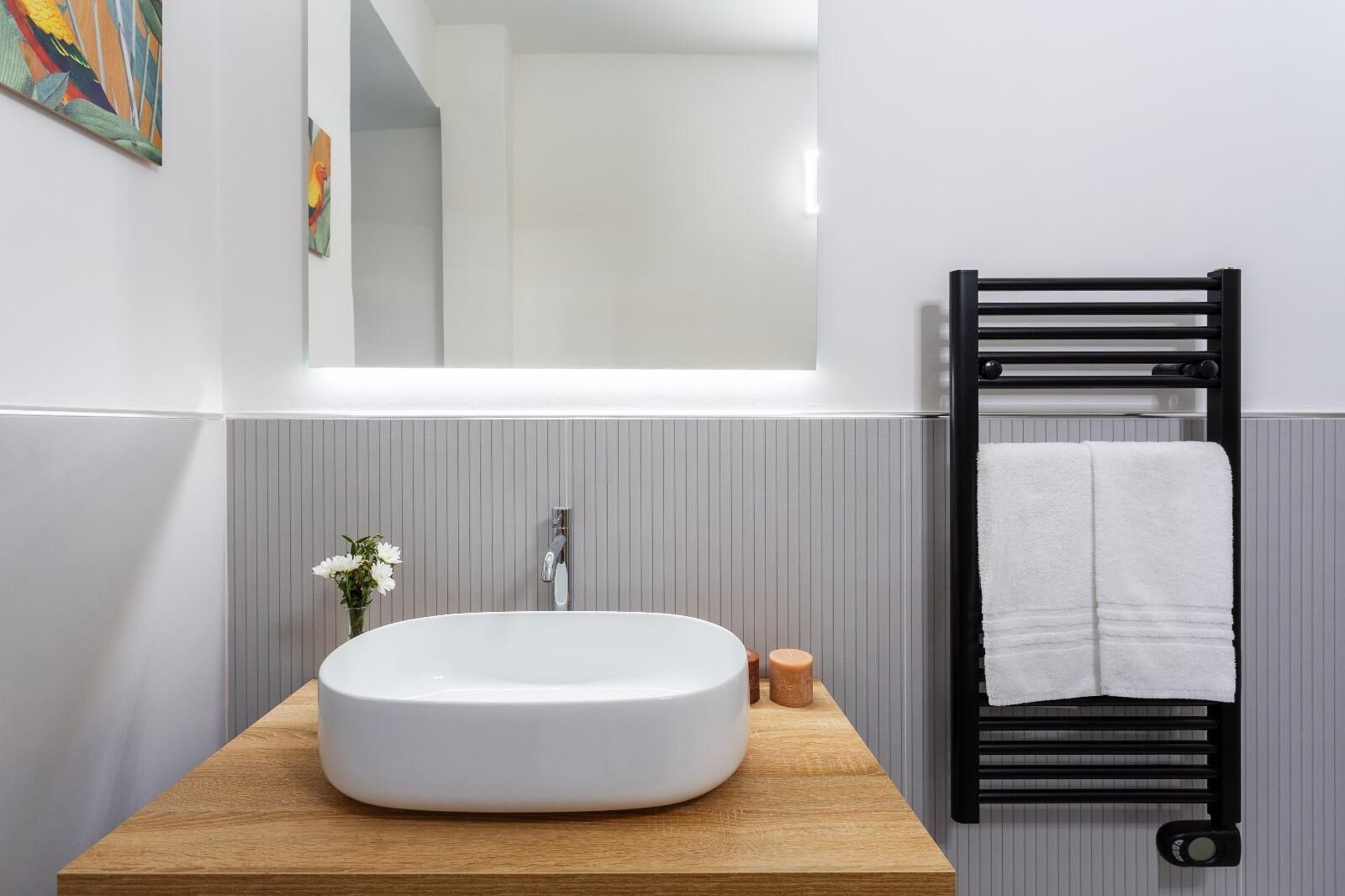 A bathroom with a round white sink, a mirror, a towel rack with a white towel, and a small vase with flowers.