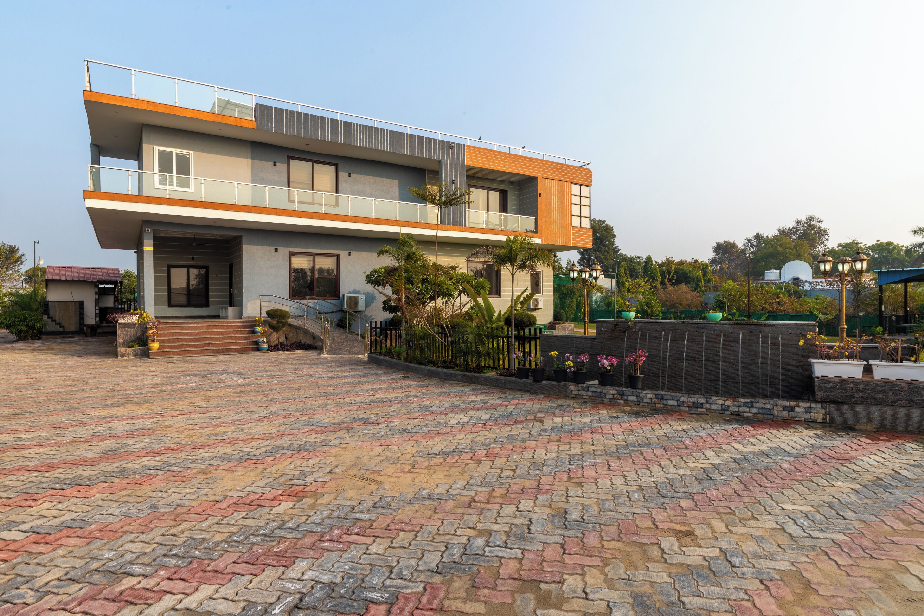 A modern two-story house with a balcony, a paved driveway, and a garden area with potted plants.