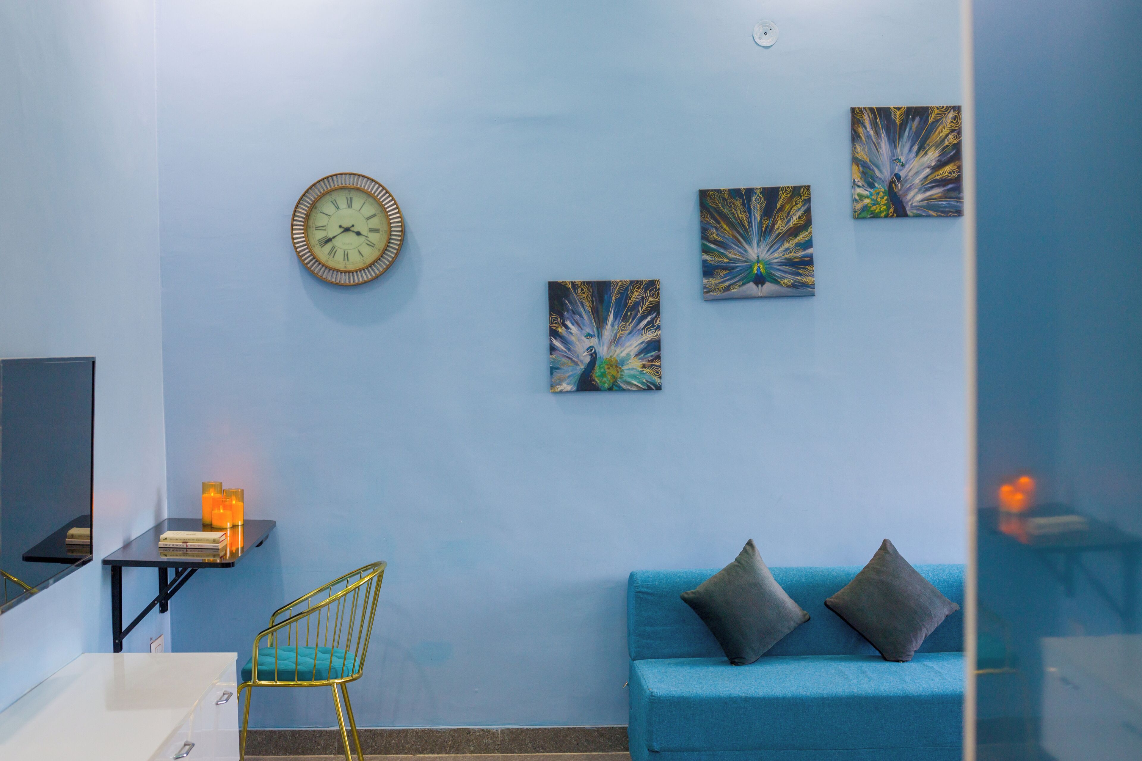Une pièce avec un mur bleu, une horloge, trois tableaux, un canapé bleu avec des coussins, une chaise jaune et une petite table avec des bougies.