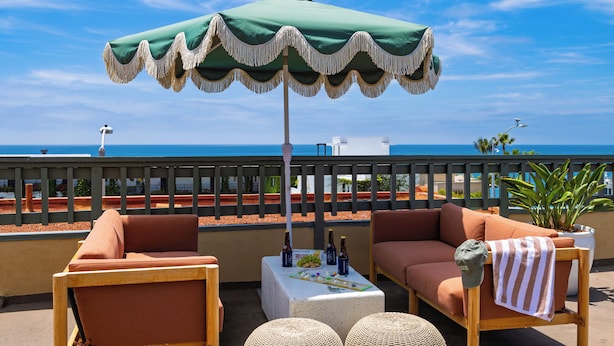 Ein Balkon mit einem grünen Sonnenschirm, zwei Sofas, einem kleinen Tisch mit Flaschen und Blick auf das Meer.