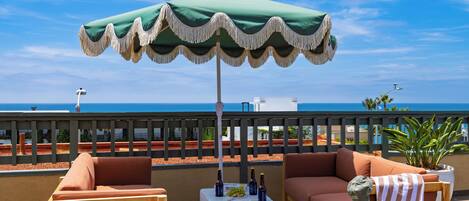 A balcony with a green umbrella, two sofas, a small table with bottles, and a view of the ocean.