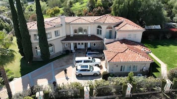 A large, multi-story house with a tiled roof, a driveway with parked cars, and a well-maintained garden.