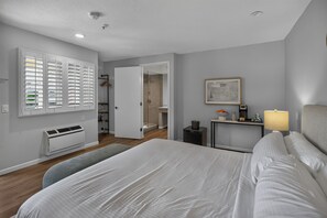 Une chambre d’hôtel comprenant un lit, une table de chevet avec une lampe, une fenêtre avec des stores, un radiateur et une porte donnant sur une salle de bain.