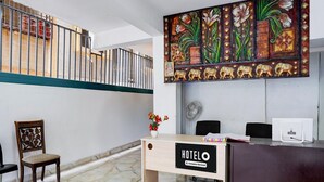A hotel reception desk with a sign, a chair, a fan, and a decorative wall piece. - Deluxesree Residency (Bengaluru)