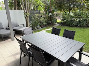 Eine Terrasse im Freien mit einem rechteckigen Tisch und vier Stühlen, umgeben von Pflanzen und einer weißen Mauer.